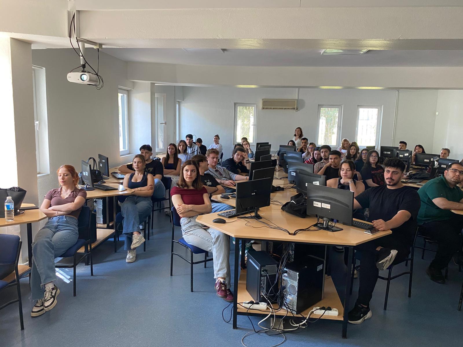In the computer laboratory, approximately 20–25 students are seated at their workstations, facing the camera. An instructor stands at the back of the room. Each workstation is equipped with a black computer. The classroom presents a bright, organized, and professional environment.